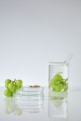A glass beaker containing fresh green grape soaked in transparent liquid and a test tube, next to a blank glass podium, decorated with some grape seed and a bunch of grape . Copy space, front view