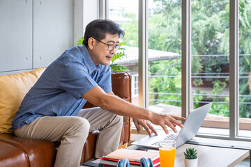 Senior Asian man using laptop at home, researching retirement plans with orange juice and notebook