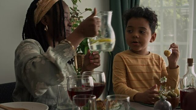 Medium shot of smiling young African American woman pouring glass of lemonade while chatting to son who eating food. Ideal for family reunion dinner commercials 