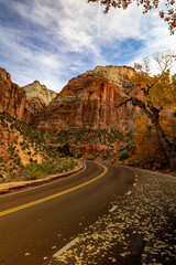 A picturesque road in Utah winds through Zion National Park. Its surrounded by red cliffs, autumn foliage, and a partly cloudy sky, perfect for outdoor adventures and exploration