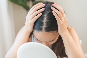 Fototapeta premium Damaged Hair symptom, face serious asian young woman, girl worry about balding, looking at scalp in mirror, hand in break into front hair loss, thin problem. Health care treatment for beauty concept.