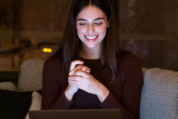 Woman sitting on sofa at night relaxes watching funny videos on her laptop. Pretty female with long hair smiles while she watches funny movie using computer connection. Dating online people. Overtime