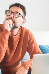 One man sitting on sofa in front of an open laptop with thoughtful expression looking for a solution or idea. People and remote working lifestyle. Small business. Security password lost problems.