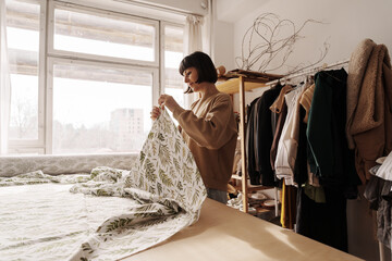 A focused artisan handles textiles in a sunlit sewing studio, surrounded by garments.