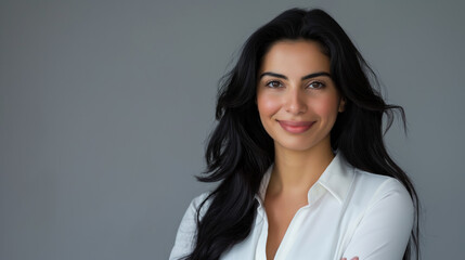 Obraz premium Portrait of a smiling businesswoman with Middle Eastern features, and long dark hair, standing with arms crossed against a light grey background