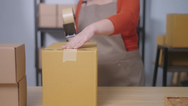 A close-up shows a woman using kraft paper tape to seal a cardboard box in a home-based business, highlighting eco-friendly packaging and sustainable practices in small business operations