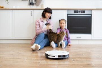 Mother and daughter playing with cat on kitchen floor while robotic vacuum cleaner operates. Modern...