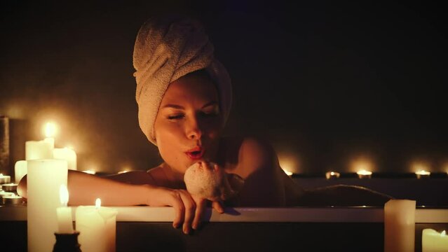Happy woman smiling face plays with soap foam in hand blowing on bulbs wet girl taking bath bubbles fly in air hair wrapped in towel enjoy bathing warm water bathtub Relax calm candles light home spa