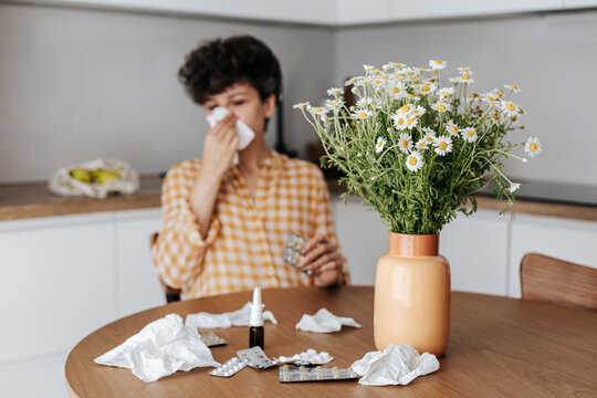 Vase of chamomile flowers on table with woman blowing nose at home