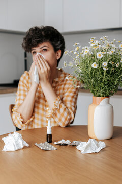 Woman blowing nose sitting near vase of chamomile flowers at home