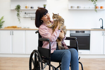 Woman in wheelchair holding pet cat in modern kitchen setting. Indoor scene showcasing bond between...