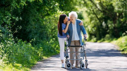 Female caregiver and senior woman with walker on walk in nature. Nurse and elderly woman enjoying a...
