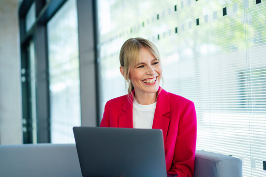 Happy blond businesswoman sitting with laptop looking away in office