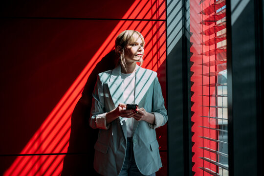 Thoughtful blond businesswoman with smart phone looking out through window at office