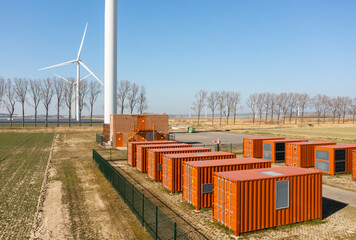 Huge red battery containers near trees and wind turbines on field