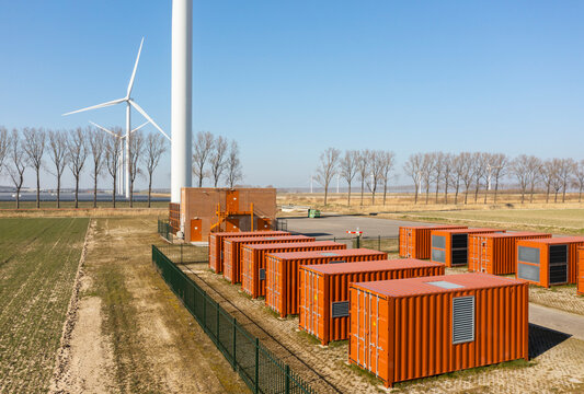 Huge red battery containers near trees and wind turbines on field