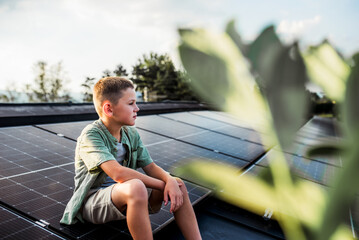 Cute boy sitting on roof with solar panels, looking at surrounding nature. Rooftop solar or...