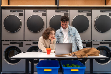 Smiling young couple using laptop on table in front of washing machines waiting at laundromat