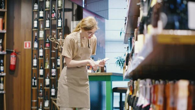 In a wine shop, an attentive female clerk is meticulously managing inventory, carefully noting details about the bottles. She appears focused and professional in her work