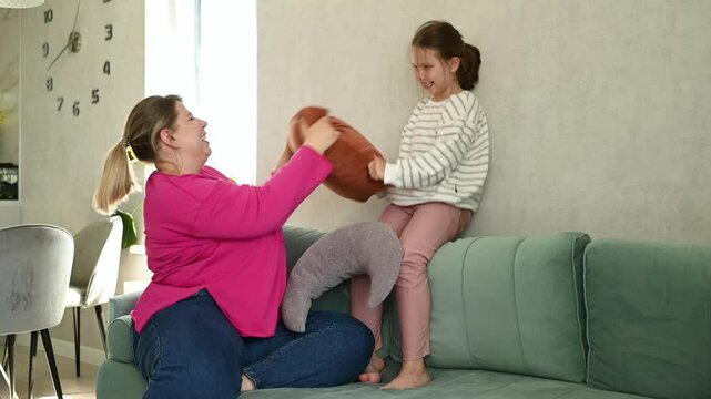 Playful mother-daughter duo having pillow fight on cozy couch