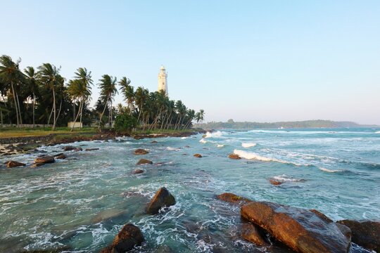A tropical rocky coast and wavy oean near the white lighthouse. Dondra Head Lighthouse is a famous landmark in Sri Lanka