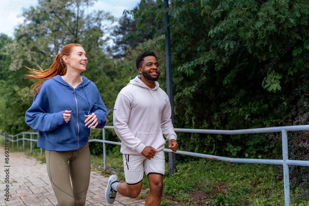 Wall mural Man and woman jogging up a hill on a paved path, laughing and enjoying their run together. They are dressed in hoodies and athletic leggings. - Wall murals