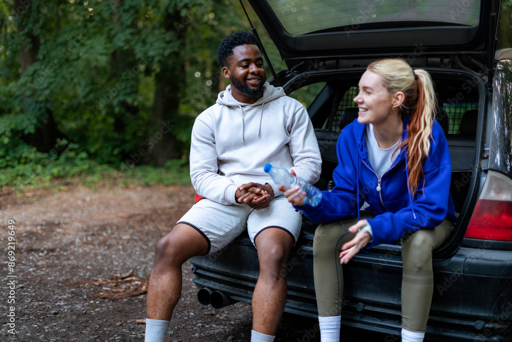 Wall mural Couple taking a water break after a run, sitting at the back of their car and chatting. They are dressed in sportswear and enjoying their outdoor workout.