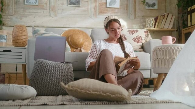 Smiling beautiful woman sitting on the floor in her cozy boho interior relaxing and enjoying reading a book
