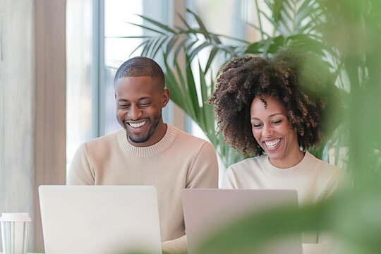 two colleagues, an African American man with short hair and a woman of mixed ethnicity with curly hair, laughing and working together on laptops in a modern office with large windows
