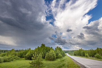 A winding country road disappears into the distance, framed by lush green trees and a dramatic, cloudy spring sky. The clouds create a sense of depth and movement, hinting at a possible storm brewing
