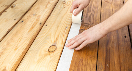 A hand carefully applies painters tape to a wooden deck, preparing the surface for a DIY project or professional painting.