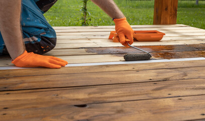 Someone in orange gloves stains a wooden deck with a roller. The deck is partially stained with...