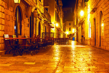 Street with tables and chairs outside of a building in the night. The street is lit up at night