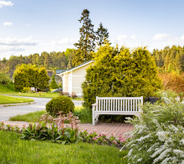 A white wooden bench sits in the center of a meticulously manicured garden, inviting relaxation beneath the shade of a tall pine tree and a vibrant yellow bush.