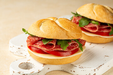 Sandwich or panino with sliced salami, sausage, tomato, cheese, rocket or arugula on a white wooden board.