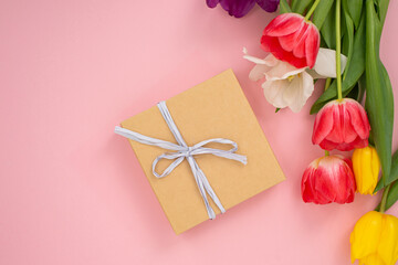 Kraft paper gift box with white ribbon next to vibrant tulips on a pink background.