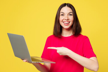 Naklejka premium Young smiling happy IT woman she wear pink t-shirt casual clothes hold use work point index finger on laptop pc computer isolated on plain yellow orange background studio portrait. Lifestyle concept.