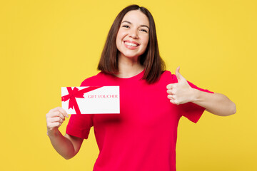 Young surprised happy woman she wear pink t-shirt casual clothes hold gift certificate coupon voucher card for store show thumb up isolated on plain yellow orange background studio. Lifestyle concept.