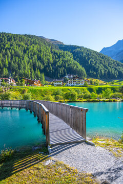 A tranquil wooden boardwalk bridge curves above the vibrant blue waters of Lake in Sulden, Italy. With the majestic Italian Alps towering in the background under a clear azure sky.