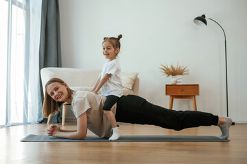 Obraz premium Kid is sitting on a back of woman. Young mother with little girl are doing yoga at home
