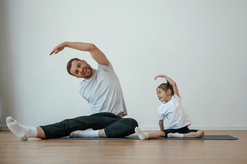 Against white wall, stretching. Father with little daughter are doing yoga at home