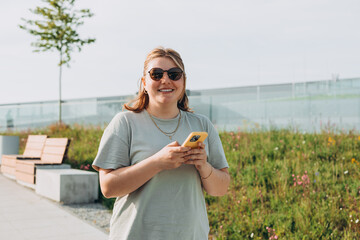 30s cheerful woman dressed in casual look holding smartphone gadget in hand for communicate on the city street. Happy woman chatting on phone and relaxing outdoors. Urban, people concept.