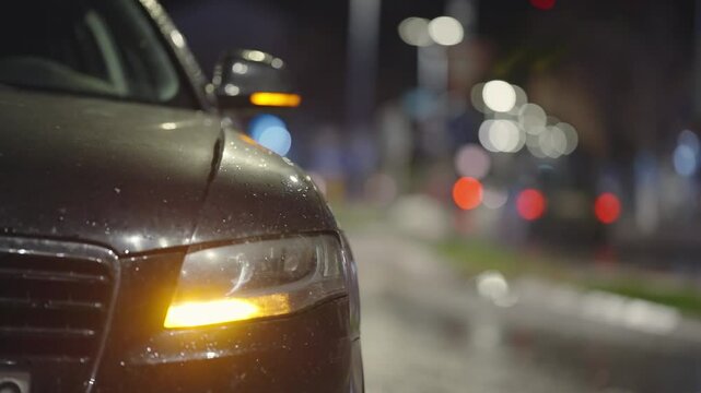Front view of parked luxury black car with flashing emergency lights and defocused city street traffic driving down a street at night. Mirror is fogged and covered with rain drops scene