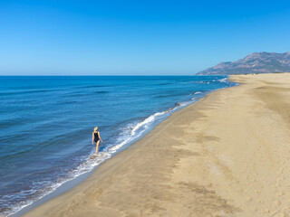 Drone view from behind young fit woman in black swimsuit running on sand along sea coast. Woman rejoices in beauty of beach, sand, nature and sea, relaxation, celebration and good mood. Joy alone.