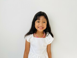 Portrait of a five year old Asian girl with shoulder length black hair wearing a white dress and necklace smiling broadly at the camera