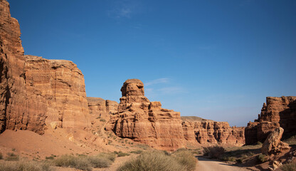 Fototapeta premium A rugged dirt road leads through an awe-inspiring canyon with towering red sandstone cliffs under a clear blue sky, highlighting the natural architecture and grandeur of the desert