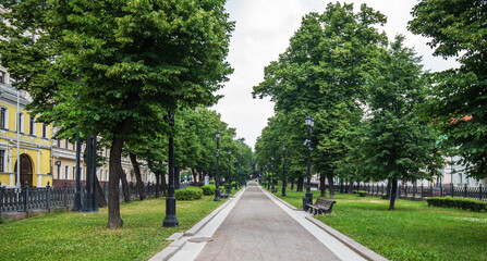 A serene, tree-lined boulevard invites a peaceful walk, with benches awaiting visitors and lamp posts standing sentinel, framing the approach to an elegant, yellow-hued building.