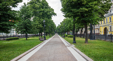 A serene, tree-lined boulevard invites a peaceful walk, with benches awaiting visitors and lamp posts standing sentinel, framing the approach to an elegant, yellow-hued building.