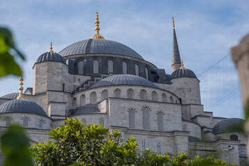Naklejka premium the timeless elegance of a mosque's domes and minarets, adorned with golden finials, set against a soft blue sky with hints of green foliage in the foreground.