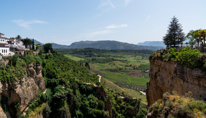 White-washed buildings crown a rugged cliff, overlooking a tapestry of green fields and distant mountains, under a bright azure sky.